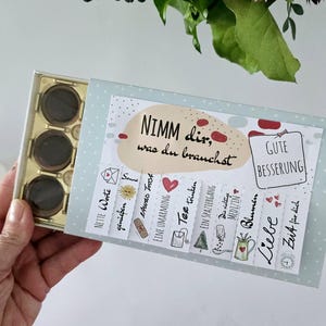 Op de afbeelding: Een geschenkdoos met een lichtblauw deksel, met chocolaatjes en een kaart met Duitse tekst. De kaart heeft verschillende illustraties en zinnen als "Goede gezondheid" en "Liefde". Een boeket bloemen staat op de achtergrond.