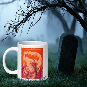Könnte beinhalten: Ein weißer Keramikbecher mit einem rechteckigen Grafikdruck auf leuchtend orangefarbenem Hintergrund. Das Design zeigt eine stilisierte, texturierte Illustration mehrerer Hände, die nach einer großen, abstrakten, zahnigen Mundform greifen.