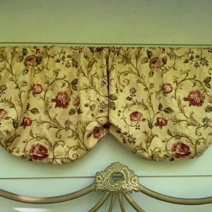 May include: A valance with a floral pattern featuring red roses and green leaves on a cream-colored background. The fabric is draped in a scalloped shape, adding a decorative touch to the window treatment. The valance is mounted above a bed frame.