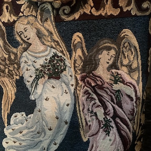 May include: A woven tapestry fabric featuring two winged angels against a dark blue background. The left angel wears a white robe and holds a bouquet of red flowers, while the right angel wears a mauve robe and holds green sprigs.