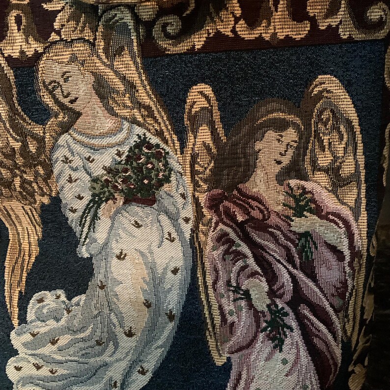 May include: A woven tapestry fabric featuring two winged angels against a dark blue background. The left angel wears a white robe and holds a bouquet of red flowers, while the right angel wears a mauve robe and holds green sprigs.