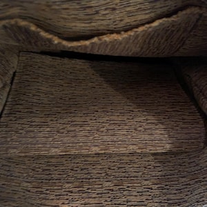 May include: The interior of a bag featuring a dark brown and tan textured fabric lining. The material displays a horizontal, irregular, and mottled pattern throughout the base and the upper pocket section, creating a consistent visual appearance.