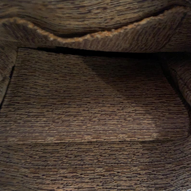 May include: The interior of a bag featuring a dark brown and tan textured fabric lining. The material displays a horizontal, irregular, and mottled pattern throughout the base and the upper pocket section, creating a consistent visual appearance.