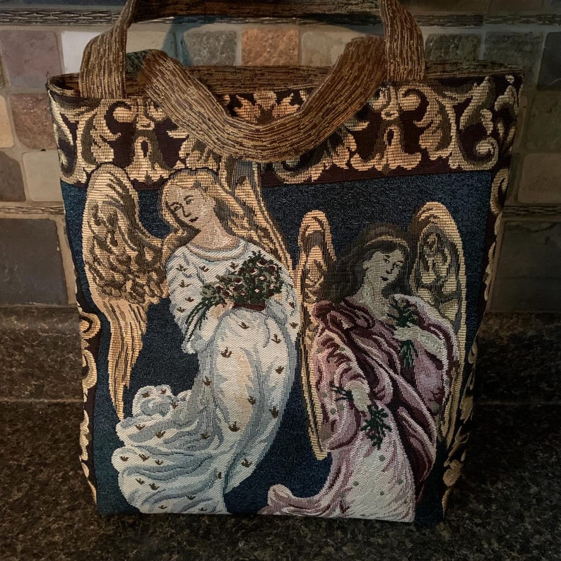 May include: A rectangular tote bag made of woven tapestry fabric featuring two winged angels against a dark blue background. The bag has brown patterned handles and a decorative border with gold and brown scrollwork designs at the top.