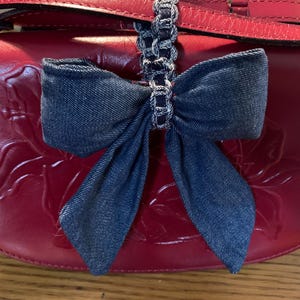 May include: A red leather saddlebag with a denim bow attached to the handle. The bag has a floral embossed design on the front. The strap is red leather and the handle is a silver chain.