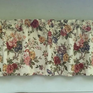 May include: A window valance featuring a cream-colored background with a dense floral pattern of roses and leaves in shades of deep red, yellow, pink, and dark blue. The fabric is gathered at the top to create soft, vertical folds across the width.