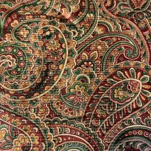 May include: Close-up of a fabric with a detailed paisley pattern. The design features swirling, teardrop-shaped motifs in shades of burgundy, green, gold, and cream. The fabric has a textured appearance, suggesting a woven material.