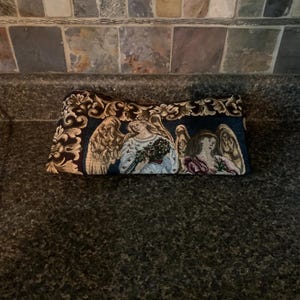 May include: A rectangular tapestry pouch featuring a woven design of two winged figures with light-colored hair and robes. The fabric displays a dark blue background with ornate gold and cream-colored floral scrollwork borders along the top edge.
