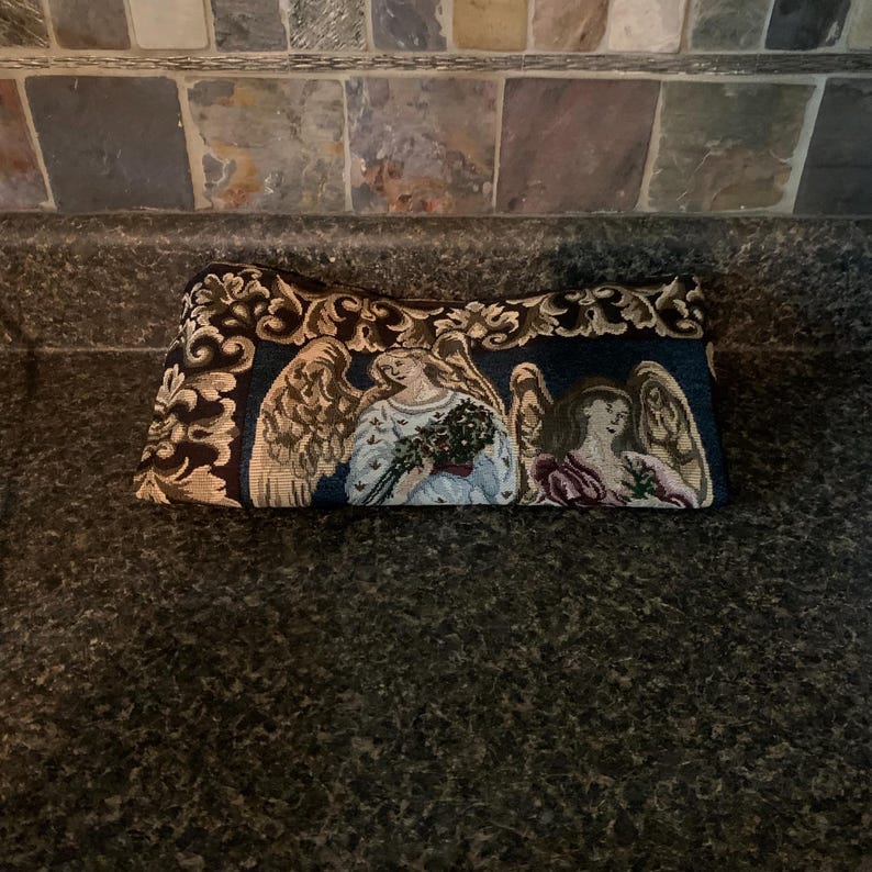 May include: A rectangular tapestry pouch featuring a woven design of two winged figures with light-colored hair and robes. The fabric displays a dark blue background with ornate gold and cream-colored floral scrollwork borders along the top edge.