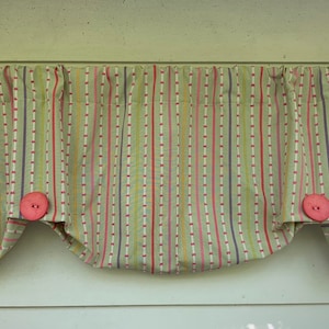 May include: A valance curtain with a striped pattern in shades of green, red, pink, and purple. The fabric is gathered at the top and held in place by two large pink buttons at the bottom corners. A decorative window treatment.