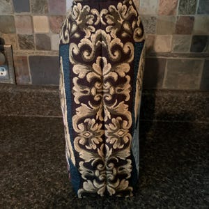 May include: A vertical fabric panel featuring a dark brown background with a prominent, symmetrical, light tan damask pattern running down the center. The sides of the panel display deep teal sections with additional intricate woven floral details.
