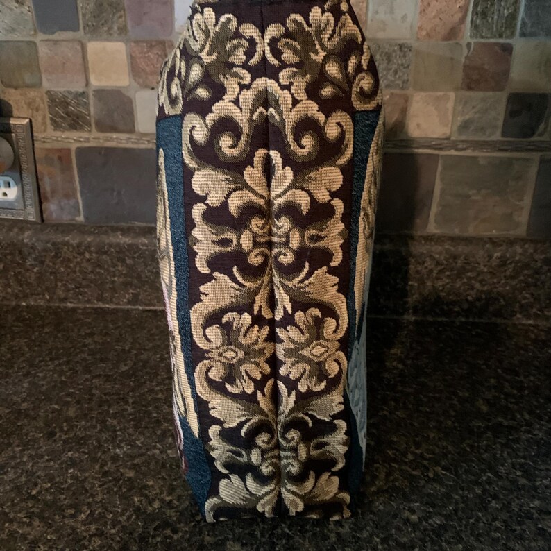 May include: A vertical fabric panel featuring a dark brown background with a prominent, symmetrical, light tan damask pattern running down the center. The sides of the panel display deep teal sections with additional intricate woven floral details.