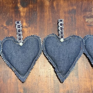 May include: Four denim heart-shaped ornaments with silver chain hangers and white pearl accents. The edges are trimmed with a decorative stitch. The ornaments are arranged in a row on a dark wood surface.