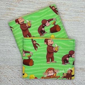 May include: Two fabric wallets featuring a bright green background with a repeating pattern of Curious George monkeys, yellow bananas, brown logs, and yellow hats. The text "Gramma's Wicked Crafty" and "Curious George" is printed on the front.