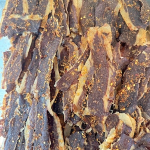 May include: Close-up of a pile of beef jerky. The jerky is dark brown with lighter-colored edges and is sprinkled with red pepper flakes. The texture appears dry and slightly rough, suggesting a savory snack.