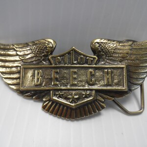 May include: A gold-toned belt buckle with a winged design. The buckle features the words "Pilot Beech" and three stars.