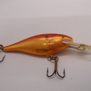 May include: A fishing lure with a gold and orange body, featuring a fish scale pattern on the top. It has a clear plastic lip and two treble hooks. The lure is designed to mimic a small fish.