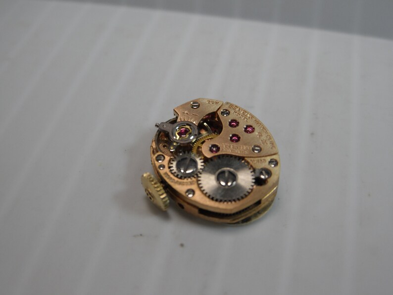 May include: Close-up of a gold-toned watch movement with visible gears and ruby jewels. The movement is labeled "Swiss Made" and "17 Jewels".