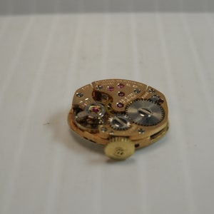 May include: Close-up of the back of a gold-toned wristwatch with a mechanical movement. The watch face is visible with a series of gears and a small red jewel. The text "MERCURY WATCH CO. SWISS MADE" is visible on the watch face.