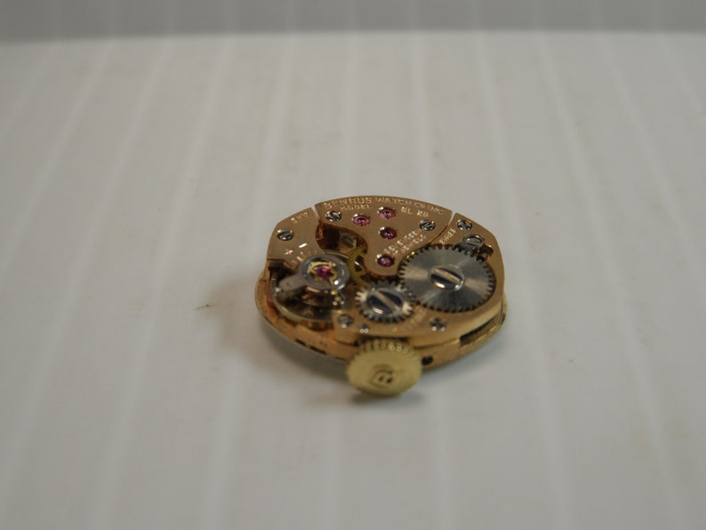 May include: Close-up of the back of a gold-toned wristwatch with a mechanical movement. The watch face is visible with a series of gears and a small red jewel. The text "MERCURY WATCH CO. SWISS MADE" is visible on the watch face.