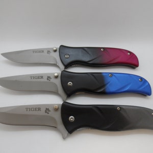 May include: Three pocket knives with black handles and silver blades. The handles have a gradient color design. The top knife has a red to black gradient, the middle knife has a blue to black gradient, and the bottom knife has a gray to black gradient. Each knife has the word "TIGER" engraved on the blade.