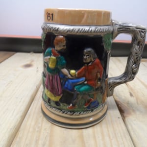 May include: A ceramic beer stein with a scene of two people in traditional clothing. The stein has a brown handle and a beige base with the number '61' on the front.
