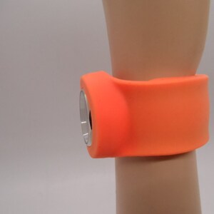 May include: Orange silicone wristband with a round, silver-colored device attached to it.