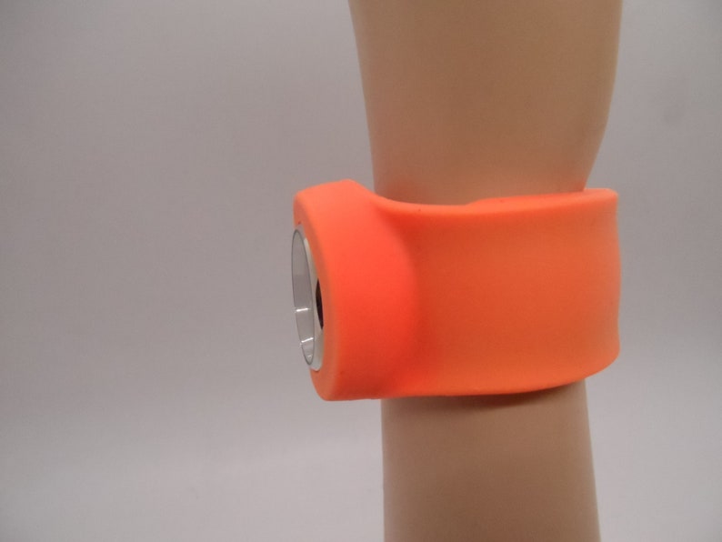 May include: Orange silicone wristband with a round, silver-colored device attached to it.