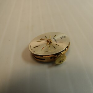 May include: A gold-toned Betrus watch movement with a white face and black hour and minute hands. The watch face has a 12 at the top and the word Betrus printed on it.