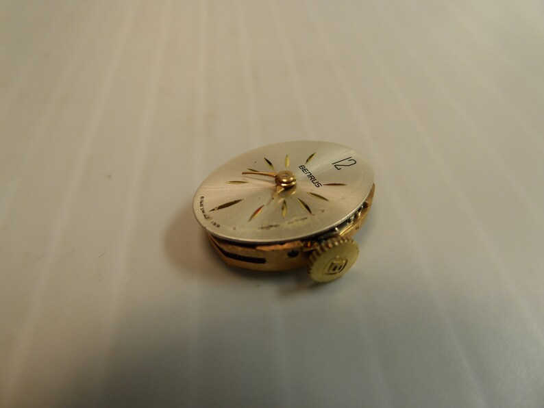 May include: A gold-toned Betrus watch movement with a white face and black hour and minute hands. The watch face has a 12 at the top and the word Betrus printed on it.