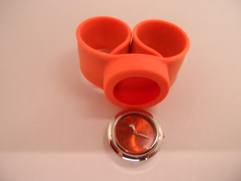 May include: Orange silicone watch holder with a silver watch face. The watch face has black hands and black numbers.