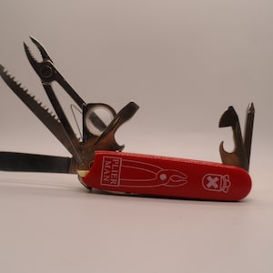 May include: A red multi-tool pocket knife with a variety of tools, including pliers, a saw, a bottle opener, and a screwdriver. The knife is closed and has the words "PLIER MAN" printed on the side.
