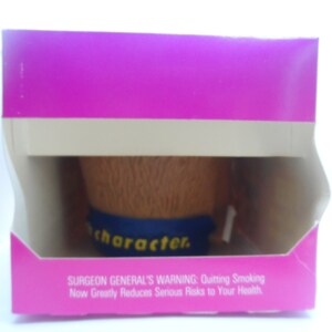 May include: A pink cardboard box with a white border and a brown and blue character figurine inside. The box has a Surgeon General's Warning about the risks of smoking.