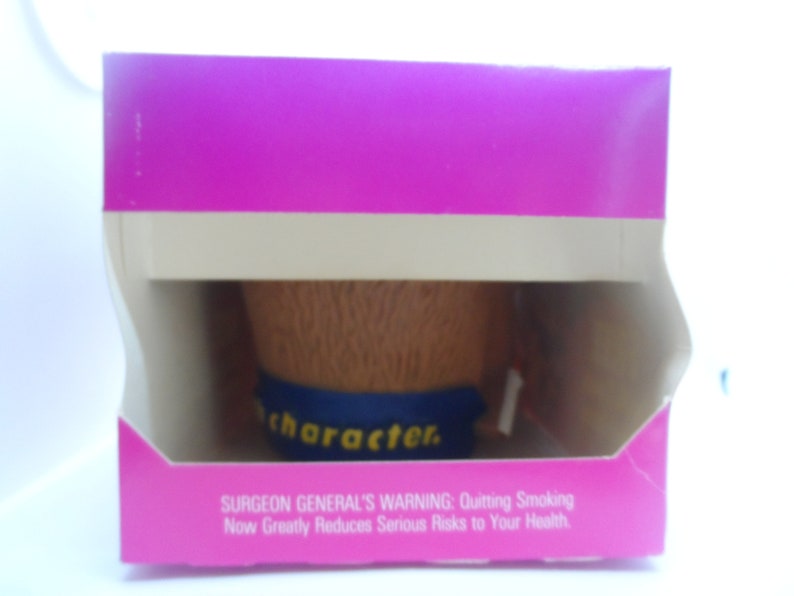 May include: A pink cardboard box with a white border and a brown and blue character figurine inside. The box has a Surgeon General's Warning about the risks of smoking.