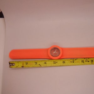 May include: A bright orange silicone wristwatch with a round face and white hands. The watch is measured against a yellow tape measure.