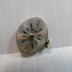 May include: A silver watch face with black markings and the word "BERNAUS" printed on it. The watch face has a 12 at the top and a winding stem on the right side.