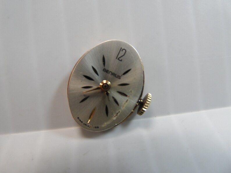 May include: A silver watch face with black markings and the word "BERNAUS" printed on it. The watch face has a 12 at the top and a winding stem on the right side.