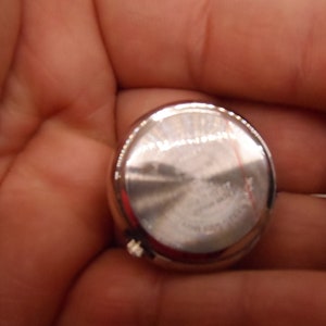 May include: The back of a silver watch with a smooth, circular case. The watch has a small, silver button on the side.
