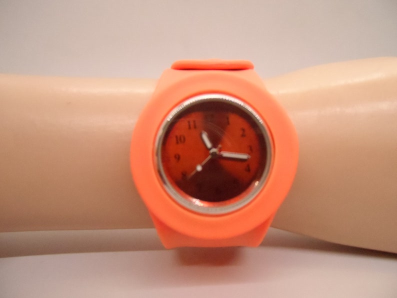 May include: A bright orange silicone wristwatch with a round face and black numbers. The watch has a silver rim around the face and a black second hand.