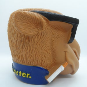 May include: A brown camel figurine wearing black sunglasses and holding a cigarette. The figurine is wearing a blue collar with the word "character" written in yellow letters.
