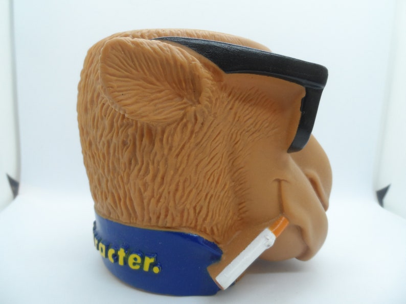 May include: A brown camel figurine wearing black sunglasses and holding a cigarette. The figurine is wearing a blue collar with the word "character" written in yellow letters.