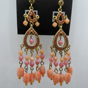 May include: A pair of gold tone dangle earrings with pink and peach colored beads. The earrings feature a teardrop shaped design with a smaller teardrop in the center.