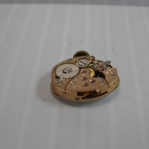 May include: Close-up of the inner workings of a gold-toned watch movement. The movement is comprised of several gears and a small, round, white dial.