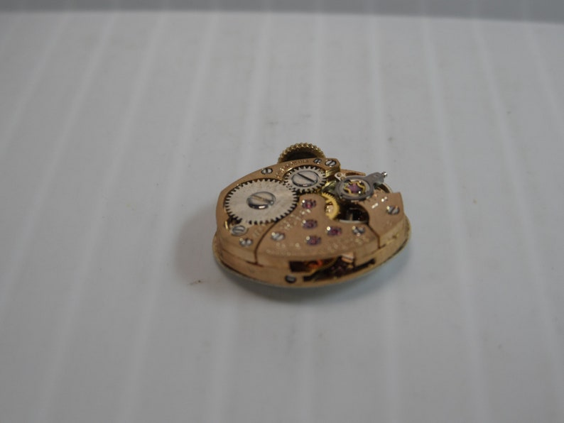 May include: Close-up of the inner workings of a gold-toned watch movement. The movement is comprised of several gears and a small, round, white dial.
