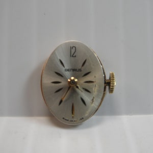 May include: A silver watch face with black markings. The watch face has a gold-colored winding stem and the text "BENRUS" and "12" printed on it.