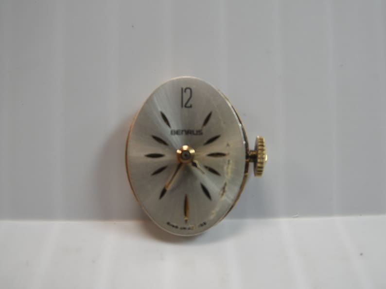 May include: A silver watch face with black markings. The watch face has a gold-colored winding stem and the text "BENRUS" and "12" printed on it.
