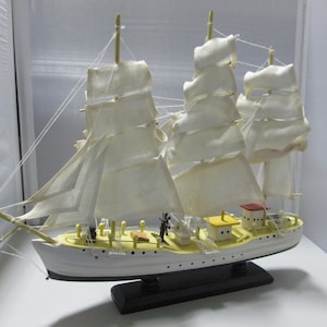 May include: A white model ship with white sails and a black base. The ship has a yellow and red deck and a wooden mast.