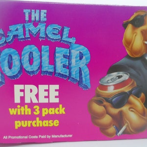 May include: A pink and blue advertisement for Camel Coolers. The ad features a cartoon camel wearing sunglasses and a suit, holding a can of soda. The text reads "The Camel Cooler FREE with 3 pack purchase. All Promotional Costs Paid by Manufacturer."