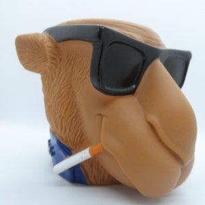 May include: A brown camel figurine wearing sunglasses and holding a cigarette. The camel has a blue bandana around its neck.
