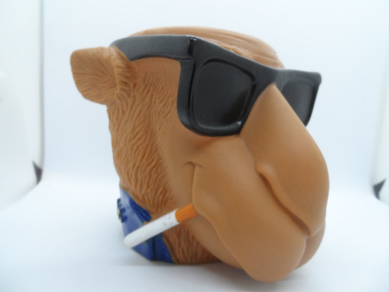 May include: A brown camel figurine wearing sunglasses and holding a cigarette. The camel has a blue bandana around its neck.